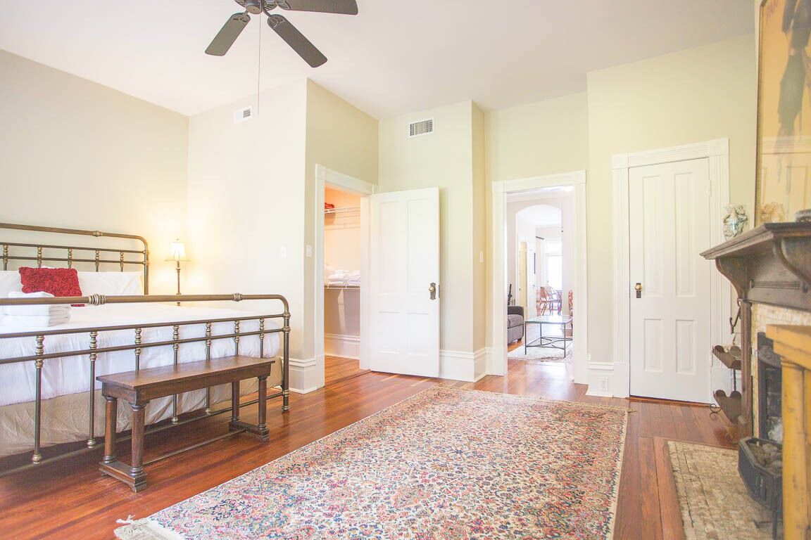 A bedroom with a bed, a bench, a ceiling fan, a fireplace, and a hallway leading to another room.