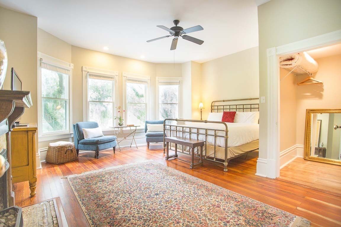 Un dormitorio con cama, ventilador de techo, ventana, silla, alfombra y cómoda.
