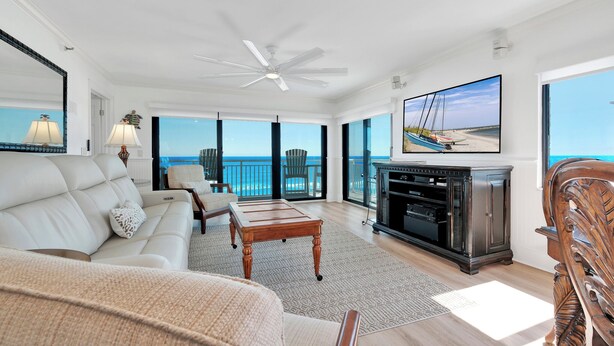 A spacious living room with a large TV, comfortable seating, a ceiling fan, and a view of the ocean. - Luxury, direct oceanfront 2/2, top-floor corner with wrap-around balcony and heated pool (New Smyrna Beach)
