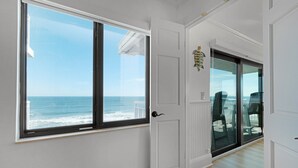A room with a large window overlooking the ocean, a white door, and a view of outdoor chairs through a glass door. - Luxury, direct oceanfront 2/2, top-floor corner with wrap-around balcony and heated pool (New Smyrna Beach)