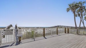 Eine Holzterrasse mit Geländer, die auf einen Strand blickt, mit Schildern zu Pool-Regeln und einem Hunde verboten-Hinweis, und einem strahlend blauen Himmel.