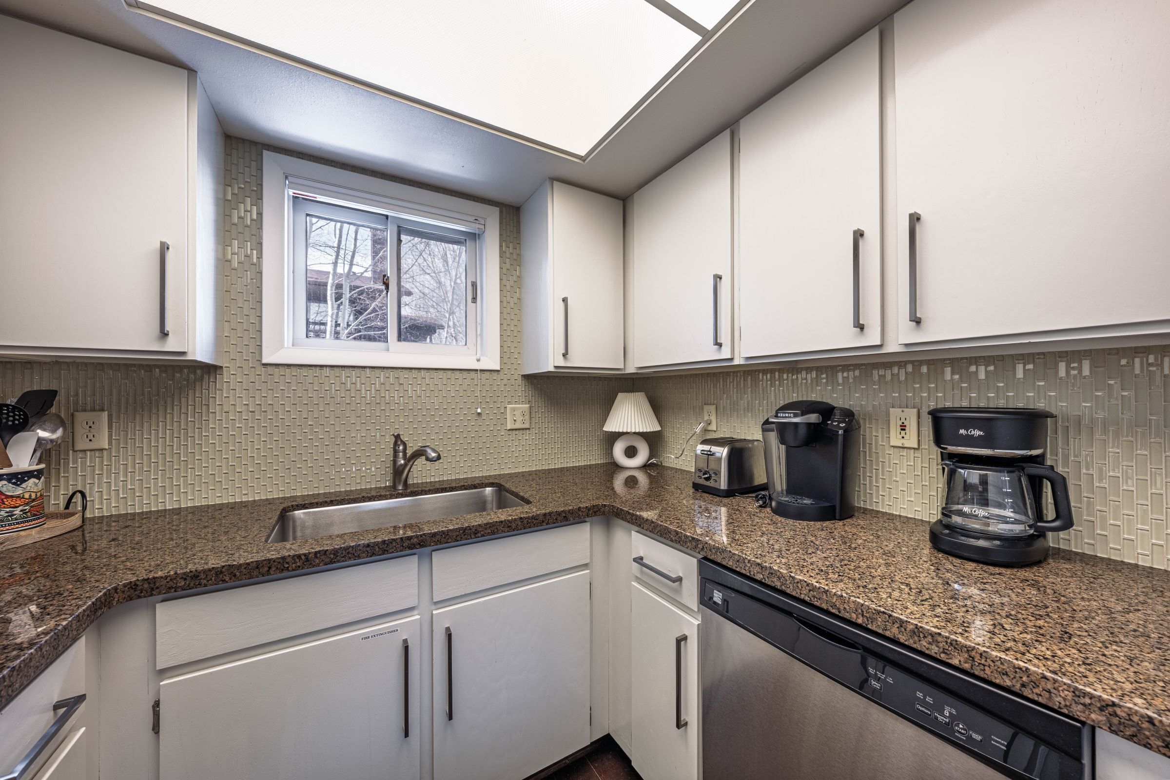 A kitchen with white cabinets, a granite countertop, a window, a coffee maker, and a dishwasher.