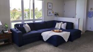 A living room with a dark blue sectional sofa, a white throw blanket, a tray with snacks, and a small table with a lamp and a plant.