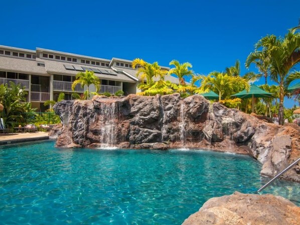 A pool with a waterfall feature, surrounded by palm trees and a building with balconies.