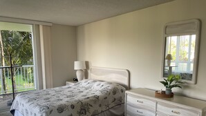 A bedroom with a bed, a nightstand, a dresser, a mirror, and a view of greenery outside.