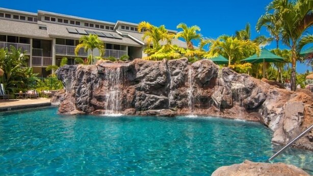 A pool with a waterfall feature, surrounded by palm trees and a building with balconies.