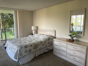 A bedroom with a bed, a nightstand, a dresser, a mirror, and a view of greenery outside.