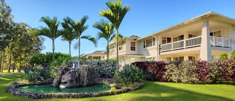 A two-story house with a balcony, surrounded by lush greenery and a small waterfall feature.