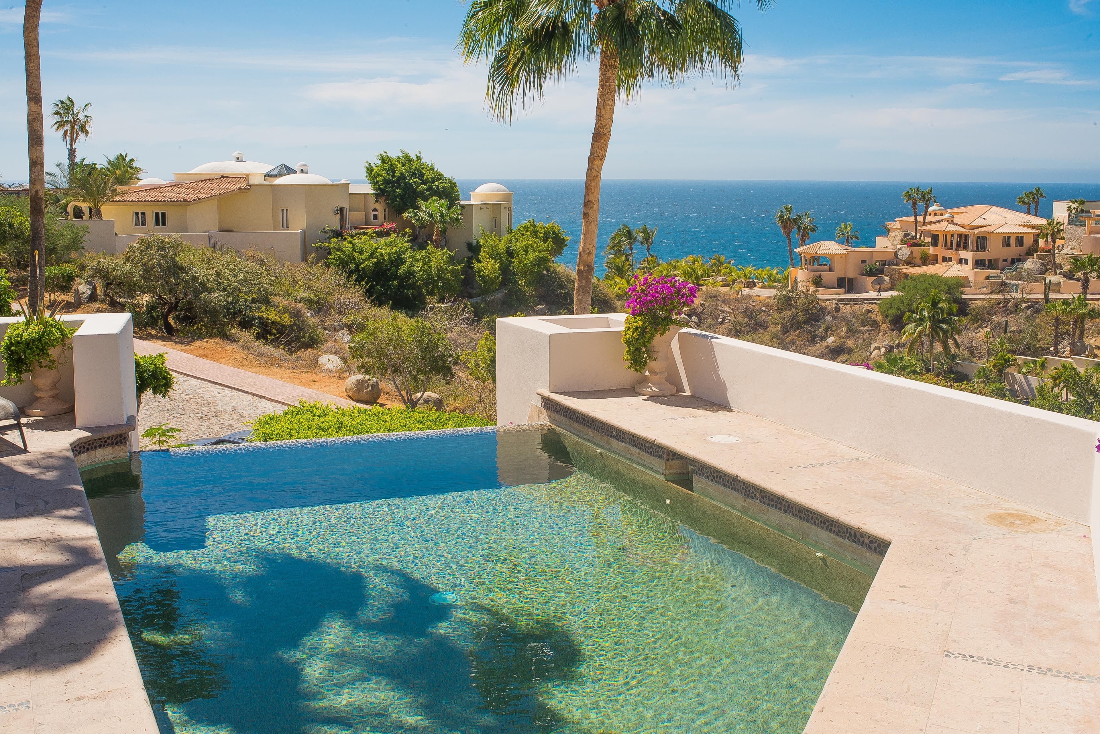 A swimming pool with clear water, surrounded by palm trees and buildings with Mediterranean architecture.