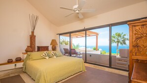 Une chambre à coucher avec un lit, un ventilateur de plafond, une grande fenêtre donnant sur l’océan et une table de chevet en bois.