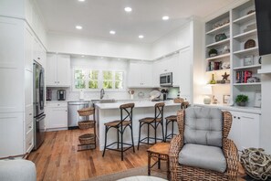 A modern kitchen with white cabinetry, a central island with bar stools, and a wicker chair. - Island House Updated DeBordieu Home just steps away from the beach (Georgetown)