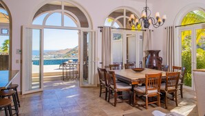 A dining area with a large table and chairs, a chandelier, and a view of the sea through open doors. - Pedregal Walk to Beach/Town Villa w/ Ocean, Bay & Marina Views, Pool & Hot Tub (Cabo San Lucas)