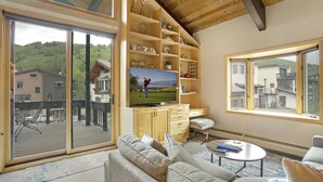 Un salon moderne doté d’un grand téléviseur, d’étagères en bois, d’un canapé et offrant une vue sur une terrasse et des immeubles.