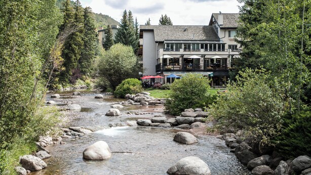Un hôtel situé au bord de la rivière, avec un sentier bordé de pierres, entouré d’arbres et offrant une vue sur les montagnes en arrière-plan.