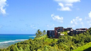 Un hôtel en bord de mer à l’architecture moderne, composé de plusieurs bâtiments aux toits plats et aux grandes fenêtres, situé sur une colline verdoyante qui surplombe l’océan.