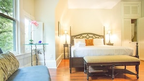 A bedroom with a large bed, bedside tables with lamps, a bench, a glass table with a vase, and a window with blinds.