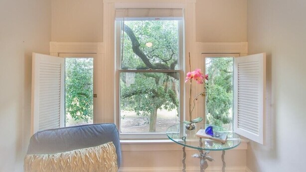 A room with a large window, a chair, and a glass table with a vase of flowers.