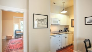A compact kitchen with white cabinets, a microwave, and a sink. There's a red rug in the hallway and a framed picture on the wall.