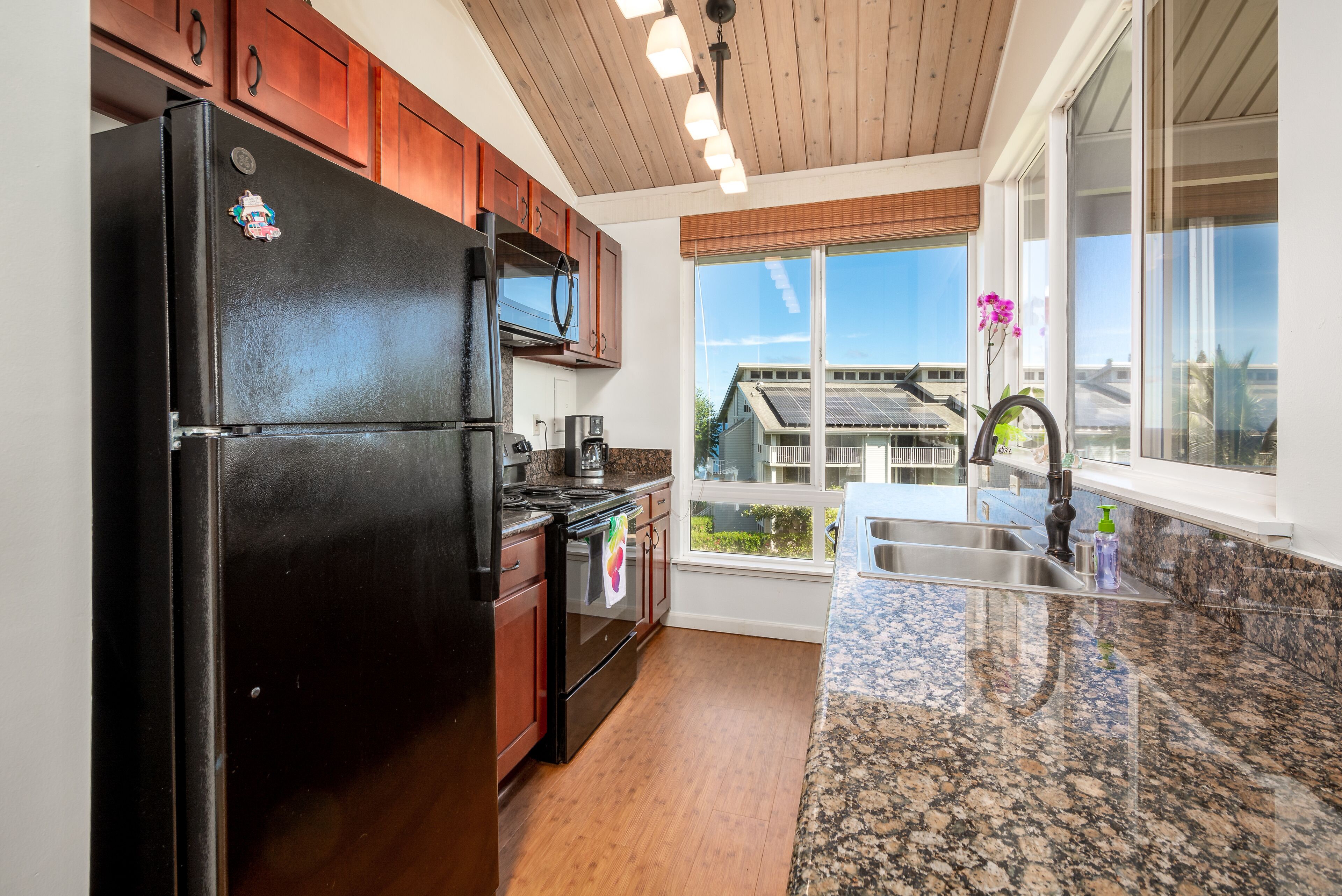 A kitchen with wooden cabinets, a black refrigerator, a microwave, a stove, and a large window with a view of solar panels and a house.