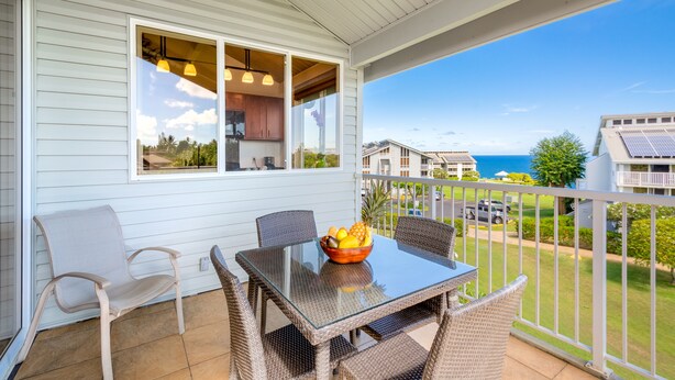 A balcony with a glass table and wicker chairs, a bowl of fruit, and a view of the ocean and neighboring houses. - Ocean and Mountain Views - Spacious Cliffs 5-301 .. 1 BR plus Loft (Princeville)