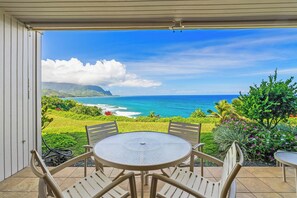 A covered patio with a round table and chairs, overlooking a scenic ocean view with lush greenery and a clear sky. - Pali Ke Kua 121 offer Stunning Oceanfront Views (Princeville)