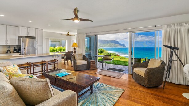 A modern living room with a large window overlooking the ocean, a kitchen with white cabinets, and a ceiling fan. - Pali Ke Kua 121 offer Stunning Oceanfront Views (Princeville)