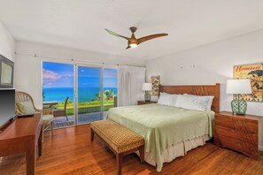 A bedroom with a bed, a desk, a bench, a nightstand, and a ceiling fan. There is a view of the ocean through the sliding glass doors. - Pali Ke Kua 121 offer Stunning Oceanfront Views (Princeville)