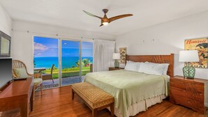 A bedroom with a bed, a desk, a bench, a nightstand, and a ceiling fan. There is a view of the ocean through the sliding glass doors. - Pali Ke Kua 121 offer Stunning Oceanfront Views (Princeville)