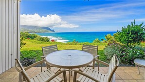 A covered patio with a round table and chairs, overlooking a scenic ocean view with lush greenery and a clear sky. - Pali Ke Kua 121 offer Stunning Oceanfront Views (Princeville)