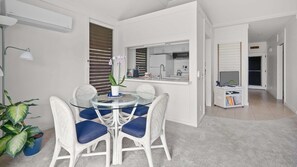 A modern kitchen with a built-in oven and microwave, a dining area with a glass table and wicker chairs, and a hallway leading to a bedroom.