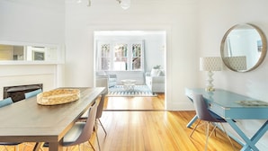 A modern dining area with a wooden table, chairs, and a woven basket. There is a blue desk with a lamp and a round mirror on the wall. - First Floor Condo w/ Large Living Area in Historic Dist. by Lucky Savannah (Savannah)