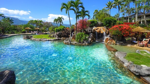 A clear pool with a waterfall feature surrounded by tropical vegetation and palm trees. - Aloha Condos, Hanalei Bay Resort, Condo 6303, Ocean View, AC (Princeville)
