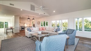 A spacious living room with a large sofa, a coffee table, and a ceiling fan. There is a kitchen area visible in the background.