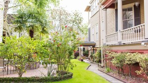 Une rue résidentielle bordée d’un jardin bien entretenu, avec une maison à deux étages dotée d’un balcon et d’un petit porche.