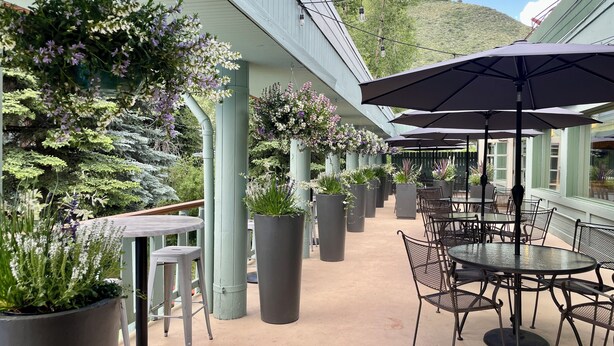 Eine Terrasse im Freien mit runden Tischen und Stühlen, umgeben von großen Kübelpflanzen und hängenden Blumenarrangements.