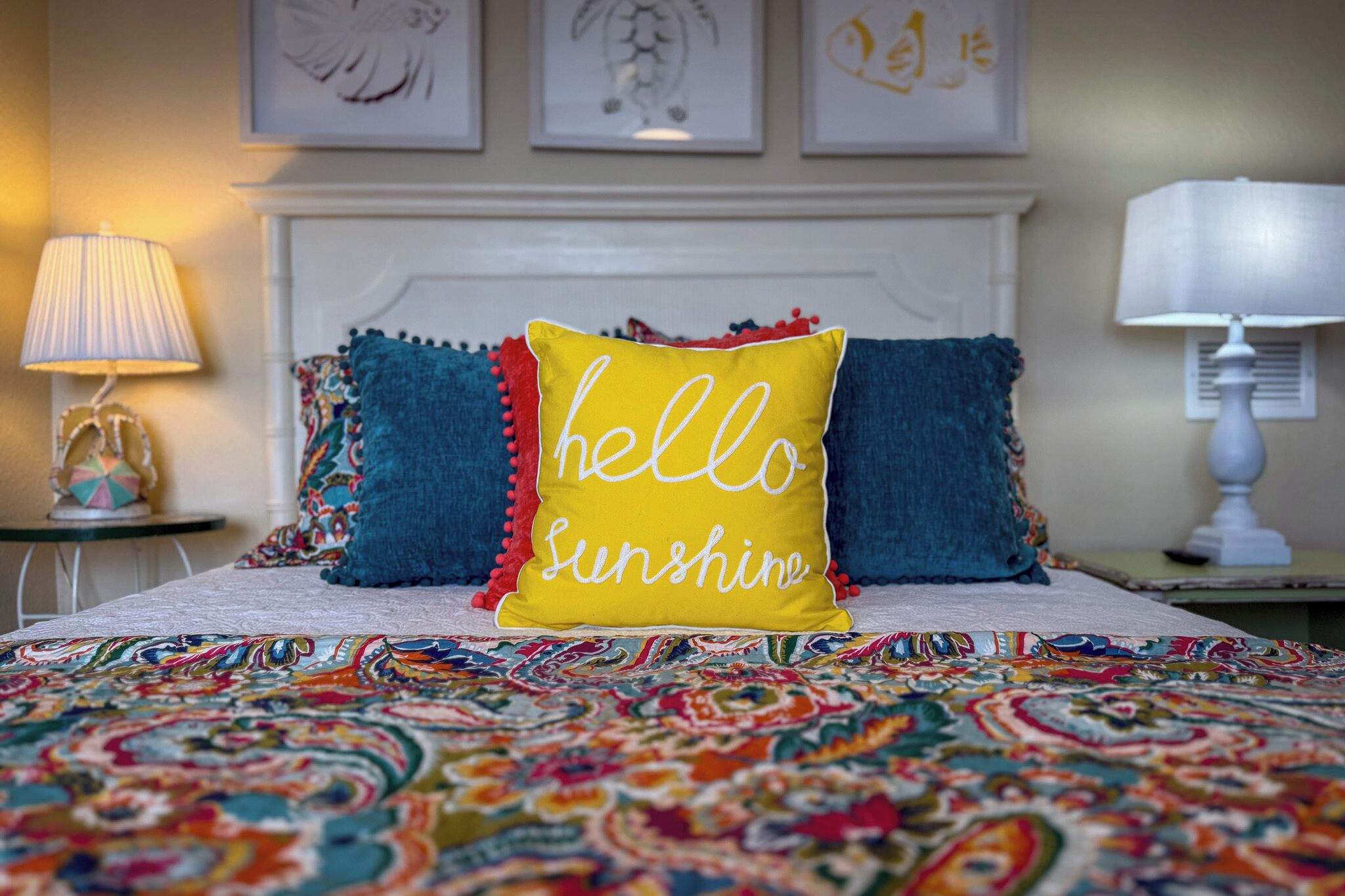 A bed with a colorful patterned bedspread, a yellow pillow with "hello Sunshine" text, and two blue pillows with tassels.