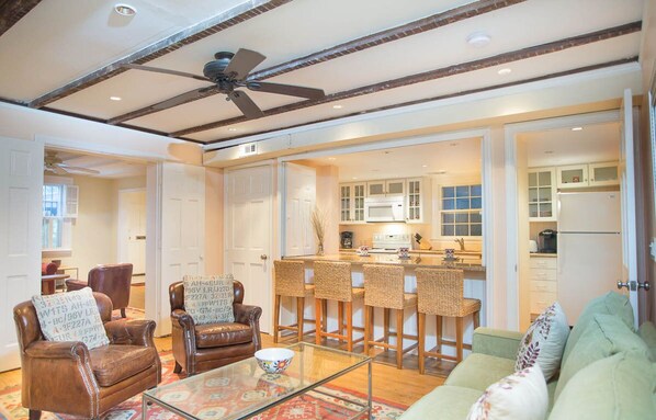A spacious living room with a high ceiling, a kitchen area with white cabinets, and a dining area with wicker chairs.