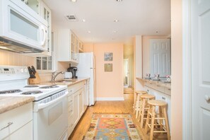 A kitchen with white cabinets, a stove, and a coffee maker. There are stools at the counter and a rug on the floor.