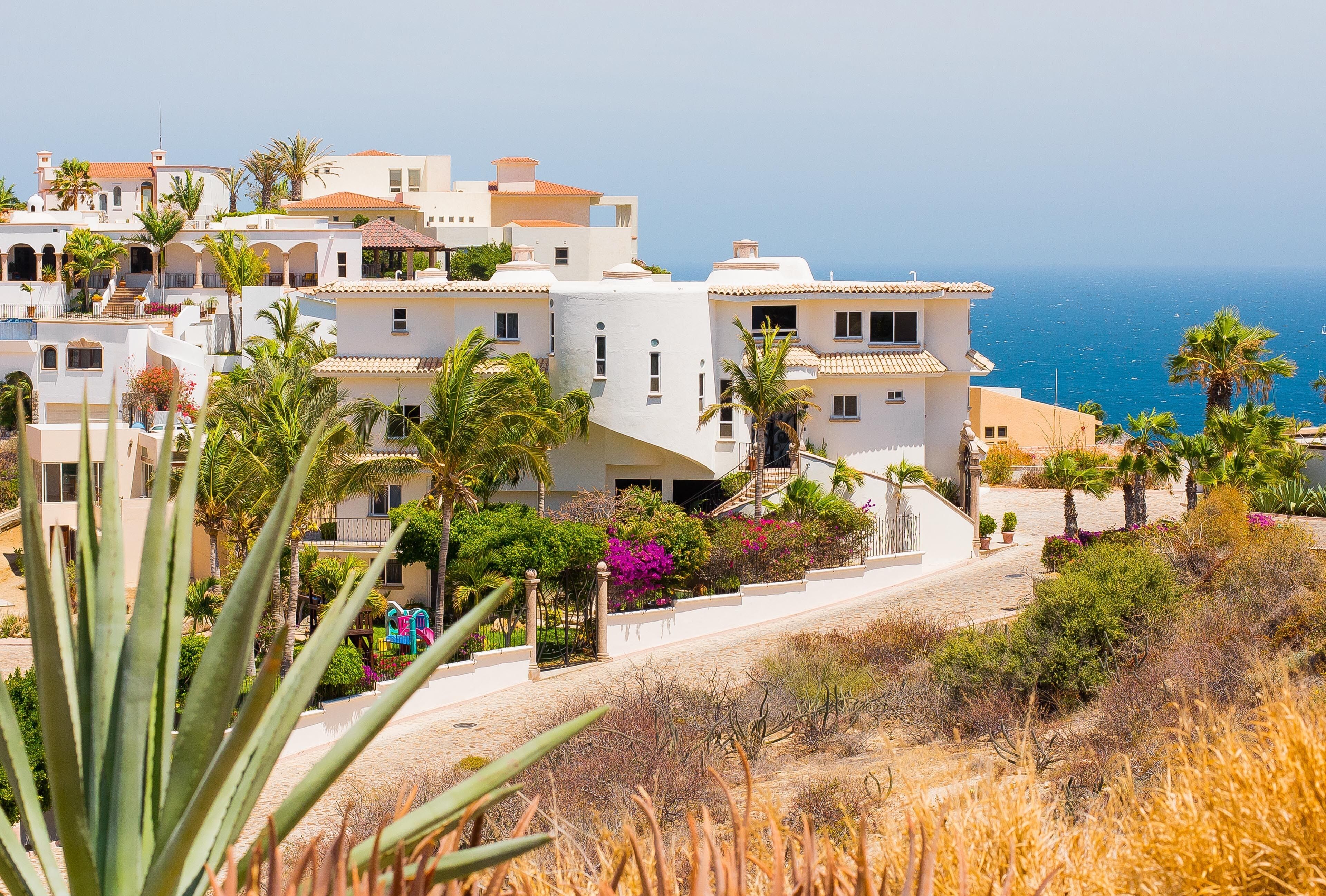 A coastal residential area with white buildings, palm trees, and a clear view of the sea.