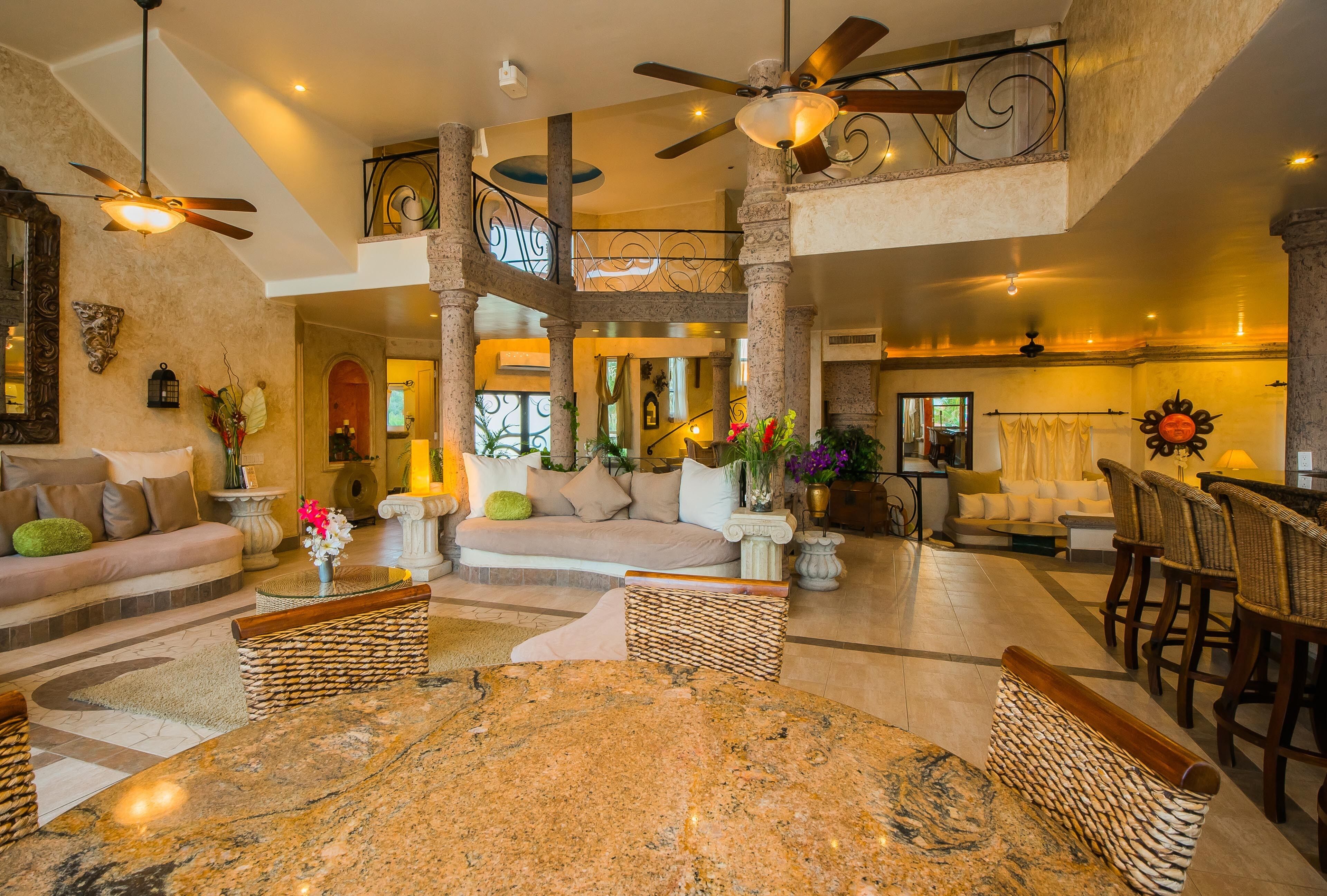 A spacious living area with a high ceiling, a central marble table, wicker furniture, and a staircase connecting two levels.