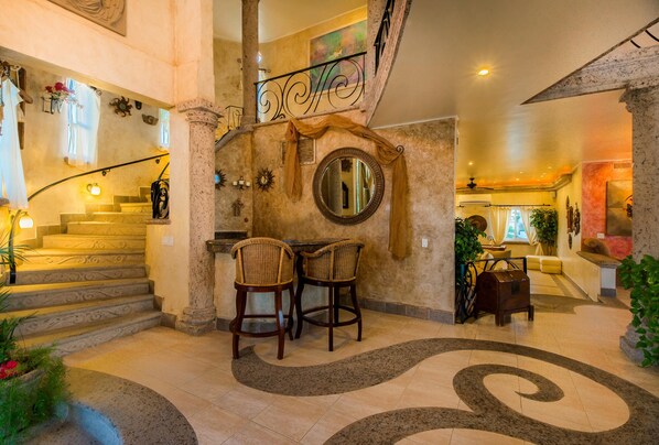 A grand staircase with a wrought iron railing, a round mirror, and two wicker chairs in a spacious lobby area. - Oceanview Villa in Pedregal Best for Groups | Pool, Hot Tub, Gym & Cabo Views (Cabo San Lucas)
