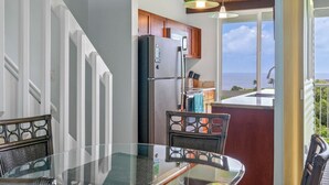 A kitchen with a view of the ocean, a dining area with a glass table, and a staircase. - Majestic Ocean View Top Floor Getaway Cliffs 7305 (Princeville)