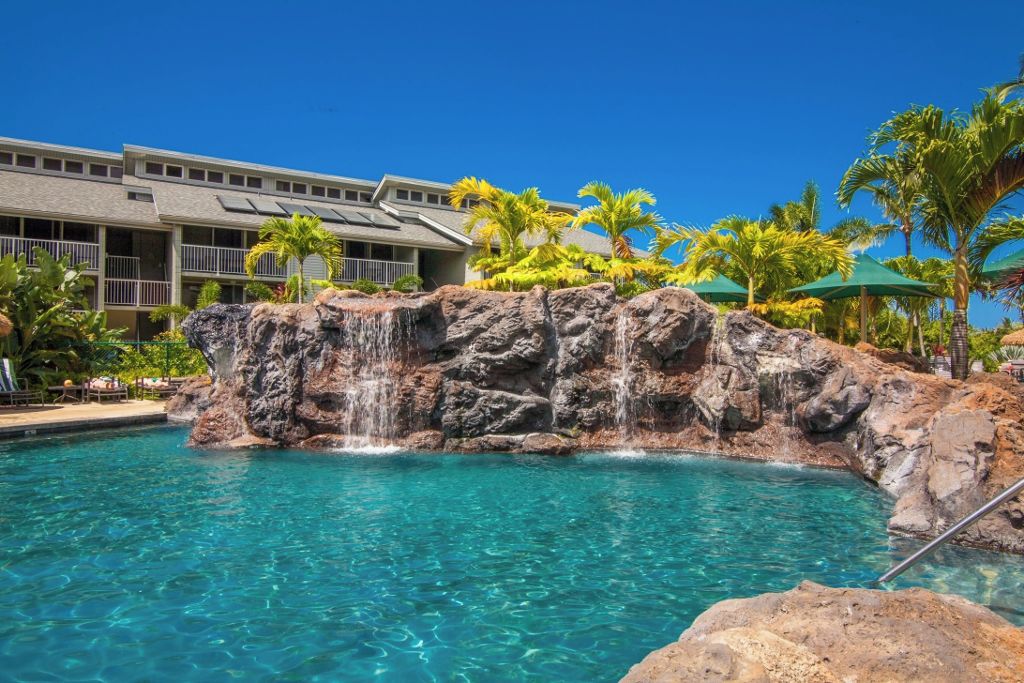 A pool with clear blue water, a rocky waterfall feature, and a building with balconies and palm trees in the background.
