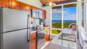 A kitchen with wooden cabinets, stainless steel appliances, and a view of the ocean through large windows. - Majestic Ocean View Top Floor Getaway Cliffs 7305 (Princeville)
