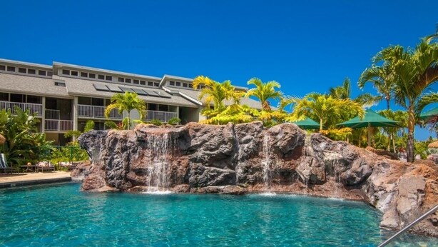 A pool with clear blue water, a rocky waterfall feature, and a building with balconies and palm trees in the background. - Majestic Ocean View Top Floor Getaway Cliffs 7305 (Princeville)