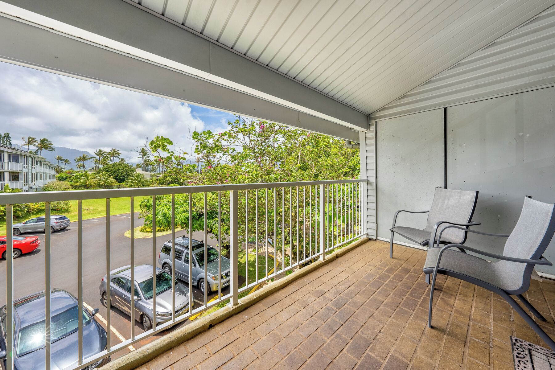 Un balcon donnant sur des voitures garées et sur de la verdure, avec une rambarde en métal et une seule chaise.