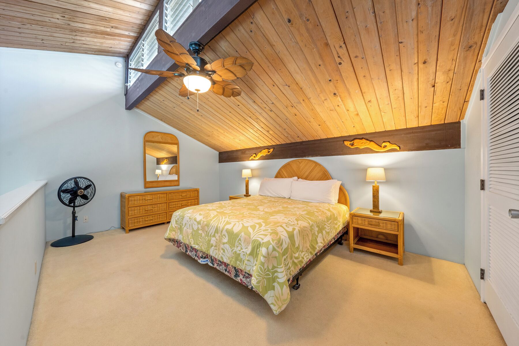 A bedroom with a sloped ceiling, a bed with floral bedding, two bedside tables with lamps, a standing fan, and a large mirror.