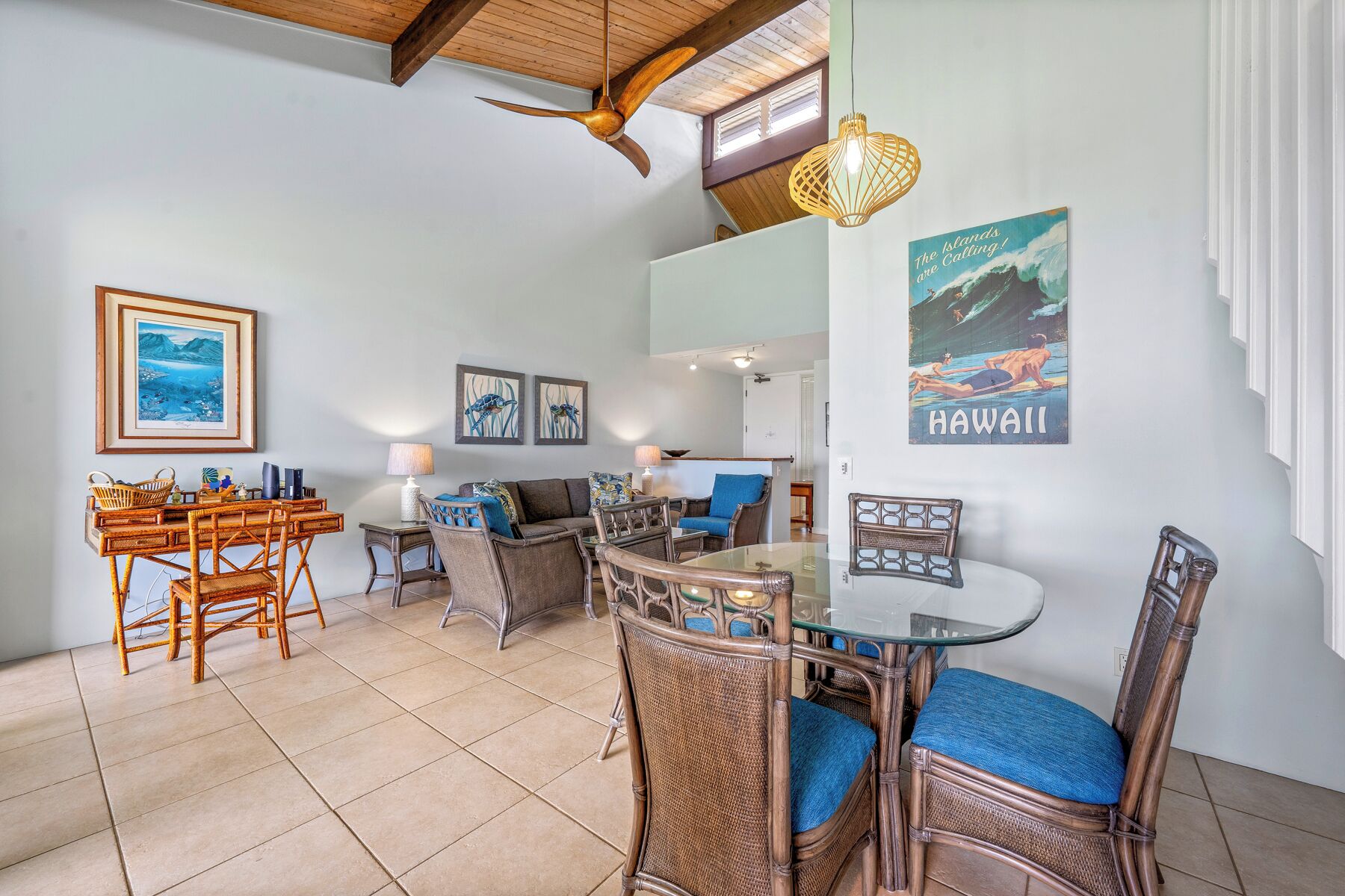 A cozy living room with a dining area, featuring wicker furniture, a ceiling fan, and artwork depicting Hawaii.