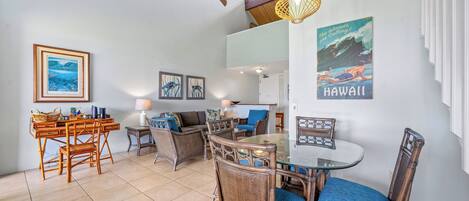 A cozy living room with a dining area, featuring wicker furniture, a ceiling fan, and artwork depicting Hawaii.