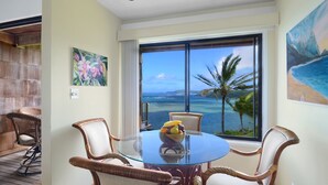 Una habitación con una mesa de vidrio, sillas de mimbre, un cuadro de flores tropicales y vista al océano.